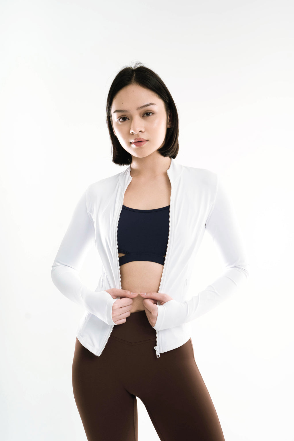 Woman wearing a white jacket over a black sports bra and brown leggings on a white background