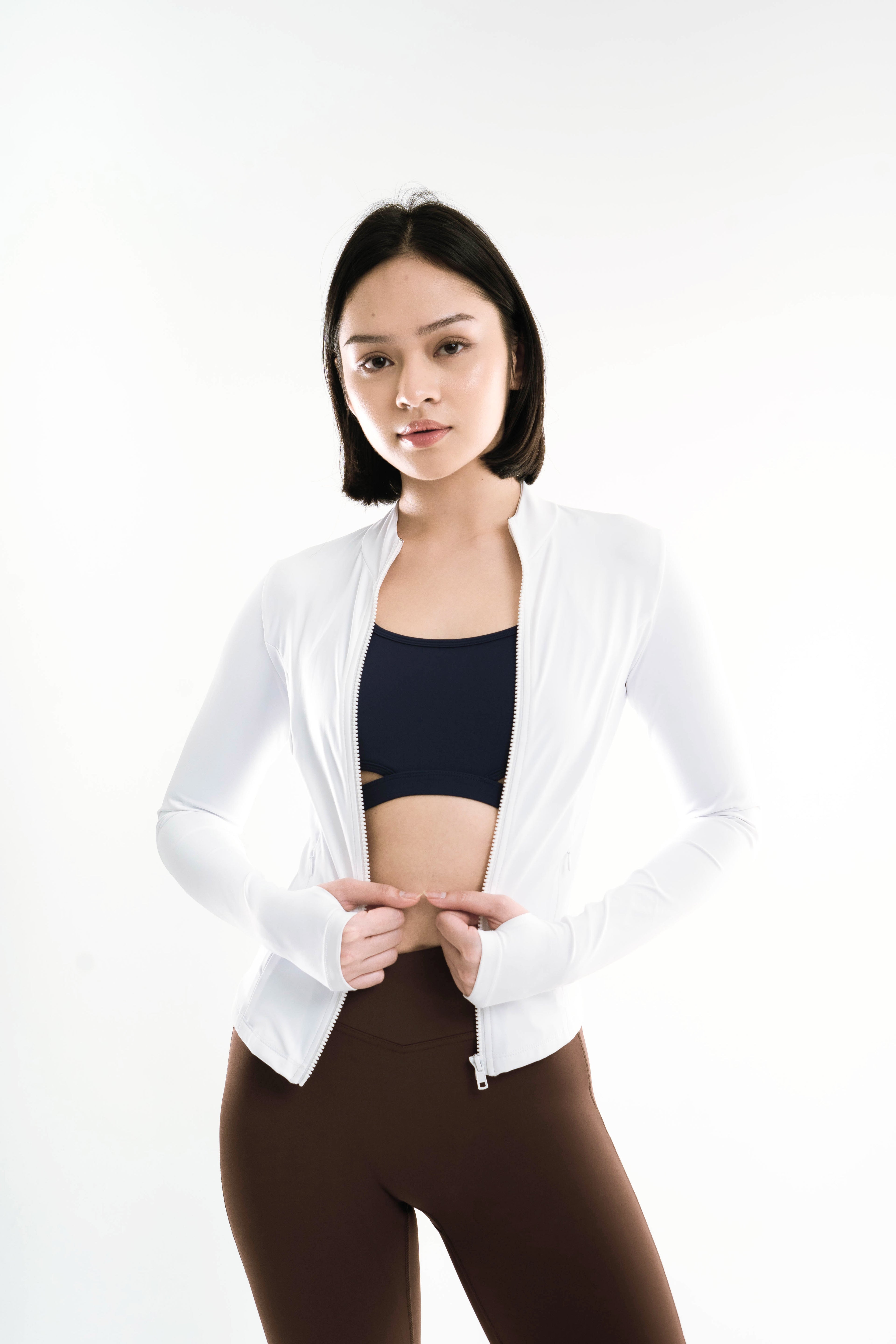 Woman wearing a white jacket over a black sports bra and brown leggings on a white background